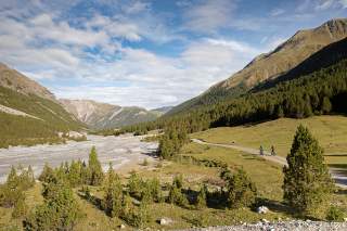 Zwei Mountainbiker fahren entlang eines breiten Kies-Flussbettes durch eine Berglandschaft.