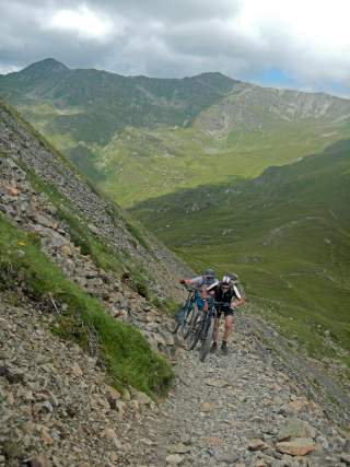 Zwei Mountainbiker schieben ihre Räder über einen schmalen Schotterweg.