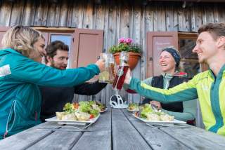 Vier Personen sitzen an einem Holztisch vor einer Berghütte und stoßen an, während auf dem Tisch Teller mit Salat und Knödeln stehen.