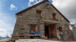 Zwei Menschen sitzen auf Bank vor Berghütte und essen.