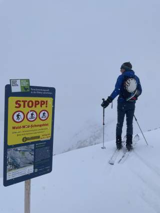 Ein Tourengeher steht im verschneiten Gelände neben einem großen Warnschild, das auf ein Wald-Wild-Schongebiet hinweist und das Betreten untersagt. Der Himmel ist wolkenverhangen, die Sicht stark eingeschränkt.