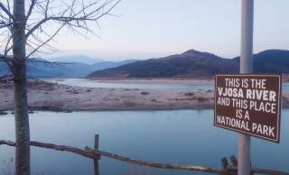 Flusslandschaft mit Bergen im Hintergrund und einem Schild am Ufer, auf dem steht: „This is the Vjosa River and this place is a national park.“