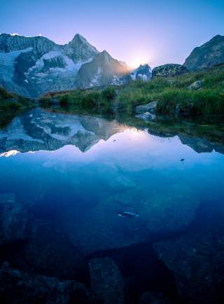 Berggipfel und die aufgehende Sonne spiegeln sich in einem Bergsee.