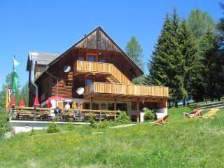 Hütte mit Balkon und Terrasse auf einer sonnigen Wiese, umgeben von grünen Bäumen, mit roten Sonnenschirmen und Liegestühlen davor.