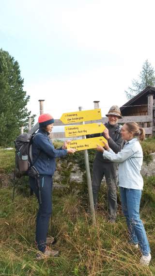Drei Wanderer stehen an einem Wegweiser mit gelben Schildern in einer bergigen Landschaft, sie halten gemeinsam die Richtungsschilder. Im Hintergrund sind Bäume und eine Holzhütte zu sehen.