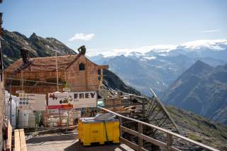 Bauarbeiter reparieren ein Holzhaus auf einer Bergbaustelle mit Blick auf schneebedeckte Berge unter klarem Himmel. Absperrungen und Baumaterialien sind im Vordergrund sichtbar.
