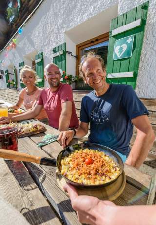 Drei Menschen sitzen auf Hüttenterrasse bei Essen und Getränken