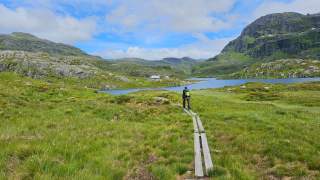 Junger Wanderer auf einem Holzbohlenweg, im Hintergrund eine Holzhütte an einem See.