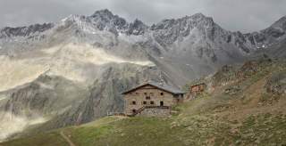 Steinernes Berghaus auf einer grünen Alm mit kahlen, felsigen Bergen im Hintergrund unter bewölktem Himmel.