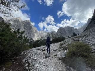 Eine Person mit Rucksack wandert auf einem steinigen Bergpfad, umgeben von hohen Felsbergen und teils bewölktem Himmel.
