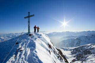 Zwei Menschen stehen bei Gipfelkreuz auf schneebedecktem Berg, die Sonne scheint