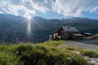 Berglandschaft mit Sonne, die hinter den Bergen herausstrahlt, und einer Berghuette aus Stein und Holz auf einer grasbewachsenen Anhöhe. Wolken ziehen am blauen Himmel vorbei.