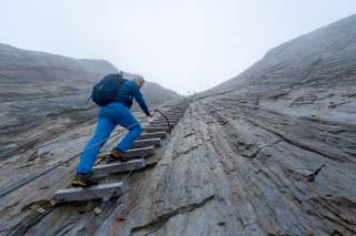 Ein Wanderer mit blauem Outfit und Rucksack klettert an einer steilen Felswand über eine Holzleiter mit Sicherungsseil nach oben. Nebel umgibt die felsige, schroffe Umgebung.