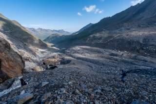 Große, steinige Gebirgslandschaft mit zerklüfteten Hängen und blauem Himmel, im Vordergrund eine einzelne Person mit Wanderstock.
