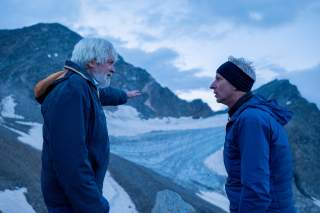 Zwei Männer in Outdoor-Bekleidung stehen vor schneebedeckten Berggipfeln, einer zeigt mit ausgestrecktem Arm in die Ferne.