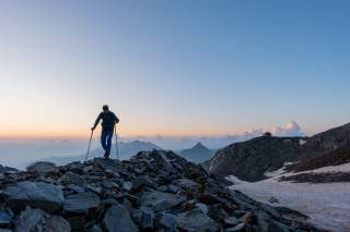 Eine Person mit Wanderstöcken steigt bei Sonnenaufgang über ein felsiges Bergplateau, im Hintergrund sind Berge und eine Hütte zu sehen.
