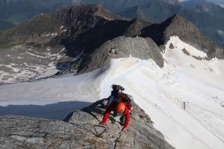 Zwei Bergsteiger klettern auf einem ausgesetzten Grat. Im Hintergrund ein Gletscherskigebiet.