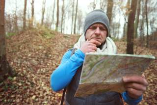 Ein Mann mit grauem Beanie und Wollschal steht im Wald und schaut rätselnd in eine topografische Karte.