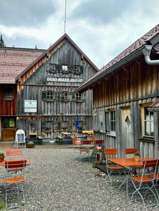 Außenbereich einer rustikalen Berghütte mit Holztischen und Bänken auf Kiesboden, das Gebäude trägt die Aufschrift „Otto Mayr Hütte“. Der Himmel ist bewölkt, und die Fassade der Hütte besteht aus verwittertem Holz.