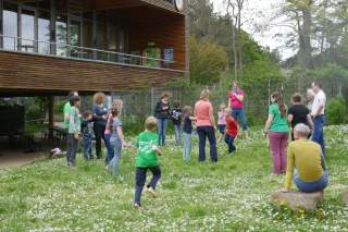 Eine Gruppe von Kindern und Erwachsenen steht im Kreis auf einer Wiese mit kleinen weißen Blumen vor einem modernen Holzhaupthaus und spielt Spiele
