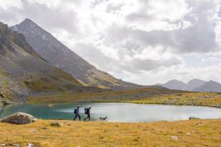 Zwei Menschen und ein Hund laufen vor Bergsee entlang