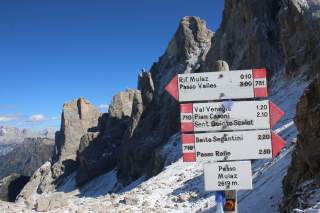 Ein rot-weißer Wanderwegweiser in den Dolomiten zeigt noch 10 Minuten bis zum Rifugio Mulaz. Hinter den Schildern ragen die Steinspitzen der Dolomiten in die Luft.