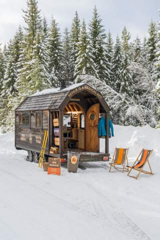 Ein Tiny House steht in verschneiter Landschaft, davor zwei orangefarbene Sonnenliegen und Ski.