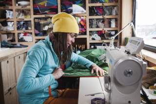 Eine Schneiderin sitzt an einer Nähmaschine und bessert ein grünes Outdoor-Kleidungsstück aus.