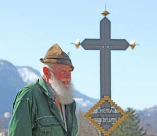 Ein älterer Mann mit grauem Bart und traditionellem Hut steht vor einem großen metallischen Kreuz mit der Aufschrift „Chiengail Kreuz“ vor einer Berglandschaft.