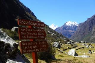 Wegweiser im Gebirge mit Hinweisen zu verschiedenen Gipfeln und deren Höhen, umgeben von Felsen und schneebedeckten Bergen unter klarem blauem Himmel.