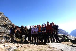 Eine Gruppe von Wanderern steht in Berglandschaft neben einem Pferd, umgeben von Felsen und schneebedeckten Gipfeln unter blauem Himmel.