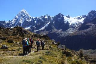 Eine Gruppe von Wanderern mit Rucksäcken und Trekkingstöcken wandert auf einem Pfad durch eine alpine Landschaft mit schneebedeckten Bergen unter klarem, blauem Himmel.