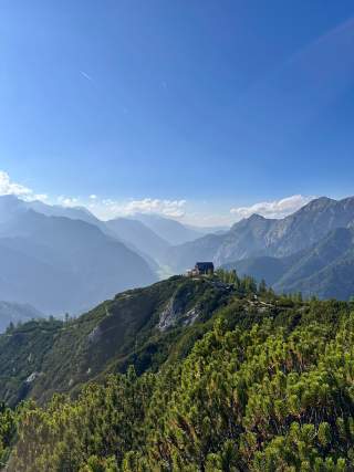 Berglandschaft mit grün bewachsenen Hügeln, einem Haus auf dem Gipfel und weiter entfernten Bergketten unter einem klaren blauen Himmel.