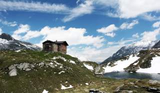 Hütte auf einer Erhöhung, rund herum Hügel und schneebedeckte Gipfel.