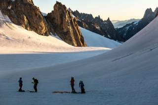 Vier Frauen in Hochgebirgslandschaft. Fels, Eis und Schnee dominieren das Landschaftsbild, ein Teil der Landschaft ist in warmes Sonnenlicht getaucht.