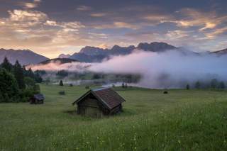Neblige Wiesenlandschaft mit Heustadeln und Bergen im Hintergrund