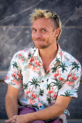 Mann mit blondem Haar und Bart trägt ein buntes Hemd mit Flamingo- und Palmenmuster und sitzt vor einer grauen Steinwand.