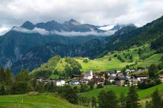 Dorf mit Kirche und mehreren Häusern in grüner Hügellandschaft vor hohen, teils bewölkten Bergen.