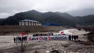 Eine Gruppe von Menschen demonstriert an einem Flussufer, umgeben von Bergen und grauem Himmel.