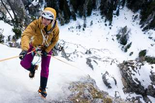 Eine Person in Winterkleidung und Helm klettert mit Steigeisen gesichert an einem eisbedeckten Felsen, umgeben von schneebedecktem Gelände und Bäumen.