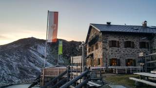 Steinernes Berghaus mit Holzbankterrasse und zwei wehenden Fahnen vor einem schneebedeckten Berg im Sonnenuntergang.