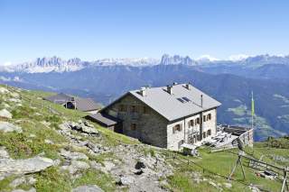 Steinerne Berghütte mit Terrasse und Sitzgelegenheiten auf einer Wiese mit Felsen, im Hintergrund hohe Berggipfel unter klarem blauem Himmel.