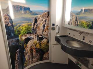 Waschbecken in einer Zugtoilette, an deren Wand ein großes Fotomotiv einer Felslandschaft mit Brücke angebracht ist.