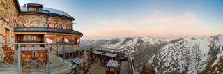 Steinernes Berghaus mit Terrasse und Holztischen, im Hintergrund schneebedeckte Alpen unter blauem Himmel bei Sonnenuntergang.