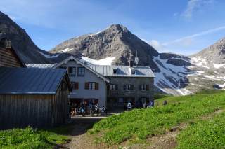 Berghütte mit Wanderern davor, umgeben von grünen Wiesen und schneebedeckten Berggipfeln unter blauem Himmel.