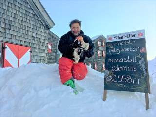 Ein Hüttenwirt mit einem Huhn vor einer Alpenvereinshütte im Schnee.