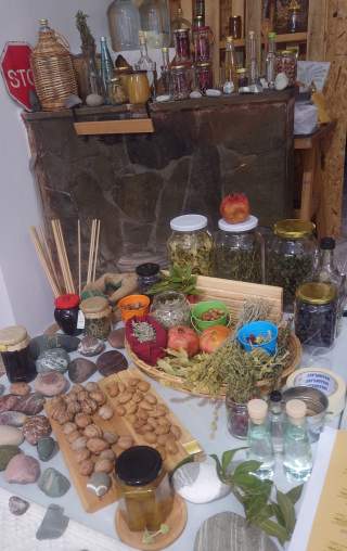 Tisch mit verschiedenen getrockneten Kräutern, Nüssen, Obst, Marmelade, Flaschen und Steinen, im Hintergrund eine Steinwand mit weiteren Gläsern und Flaschen.