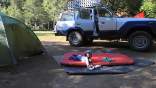 Nelion liegt auf einer Luftmatratze, im Hintergrund der Geländewagen und ein Zelt.