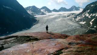 Eine Wanderin steht klein vor einem großen Gletscher, der sich (noch) zwischen den Bergen durchschlängelt.