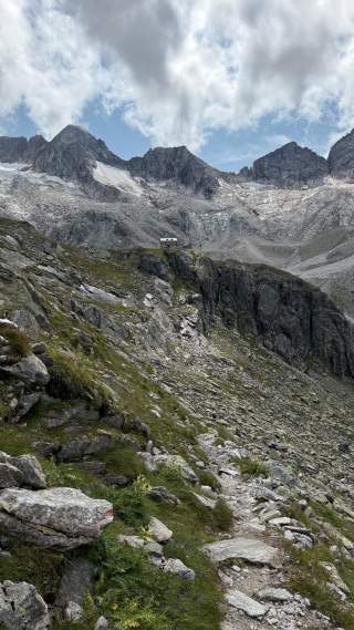 Wanderweg führt über felsiges Gelände und grüne Vegetation zu einer Berghütte vor schroffen Gipfeln unter bewölktem Himmel.